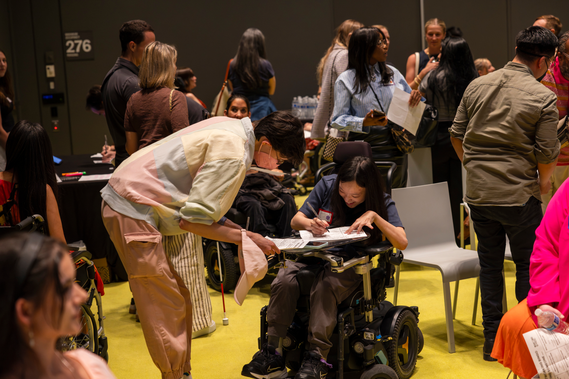 An attendee leans down to help another participant in a power wheelchair fill out a networking card.